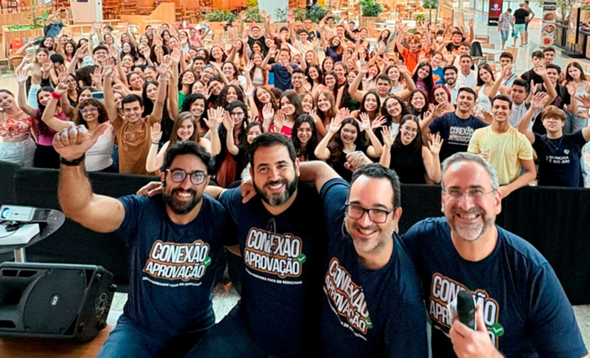 Quatro professores sorrindo para a câmera e várias pessoas com as mãos levantadas ao fundo